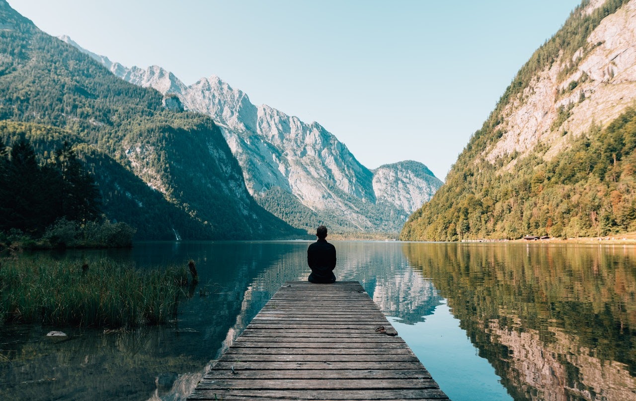 A man sits on a bridge looking out at a mountain landscape. He appears to be tranquil and in a peaceful state.