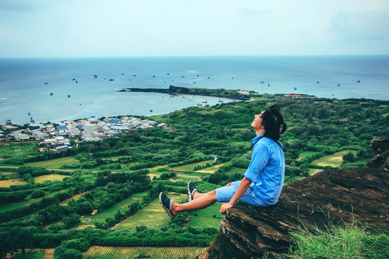 Man Smiles Happily Over Grassy Fields While Sitting On Cliff