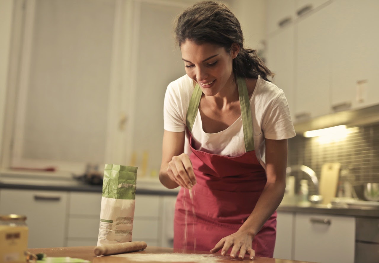 A woman smiles while sprinkling flour on kitchen counter. The woman wears an apron and loves cooking as a hobby.