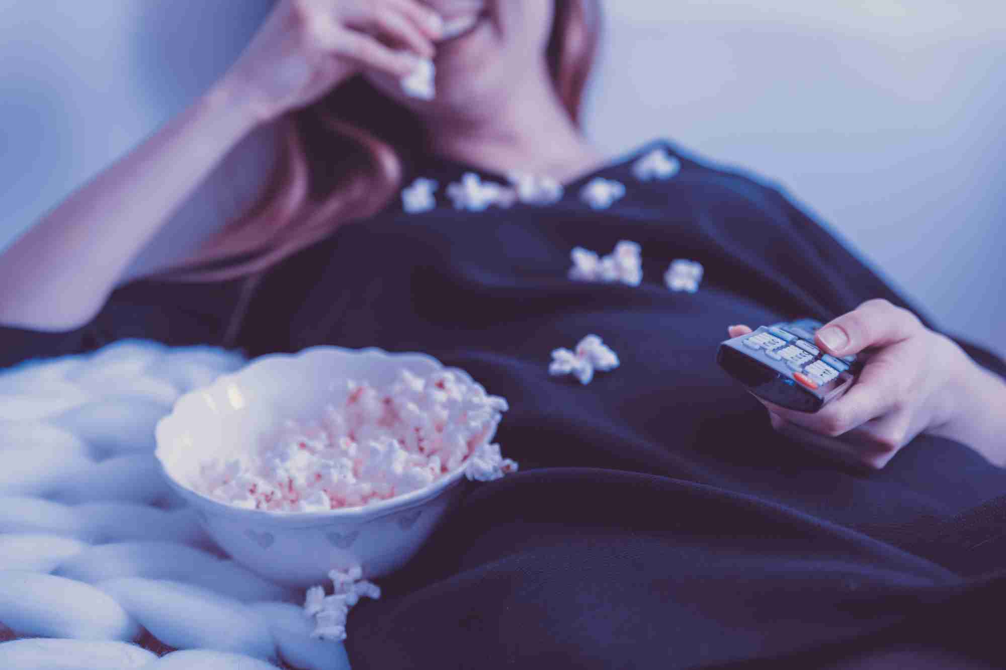 Mujer con pijama tumbada cómodamente en un sofá, con el mando de la televisión en una mano, comiendo palomitas de maíz con la otra.