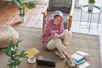 a young woman sitting on a floor with headphones on, with a coffee, a laptop, books and notes around