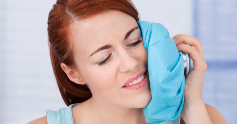 A woman holds a blue ice pack to the left side of her face to help reduce pain and swelling.