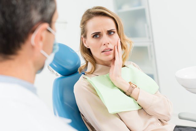 A woman in a dentist's chair holds her face and looks at a dentist with his back to the camera.s