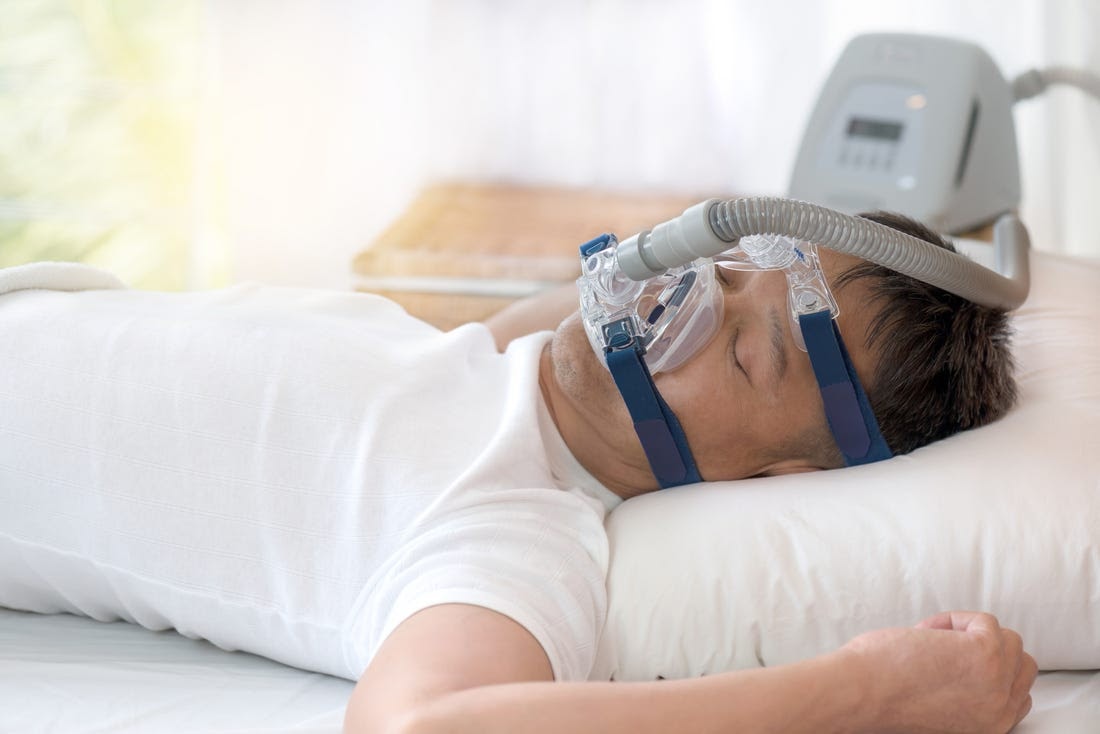 A man in a white shirt uses a CPAP machine for sleep apnea.