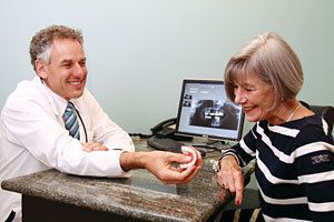 Dr. Burden sits behind desk and hands denture to woman in a black-and-white striped shirt