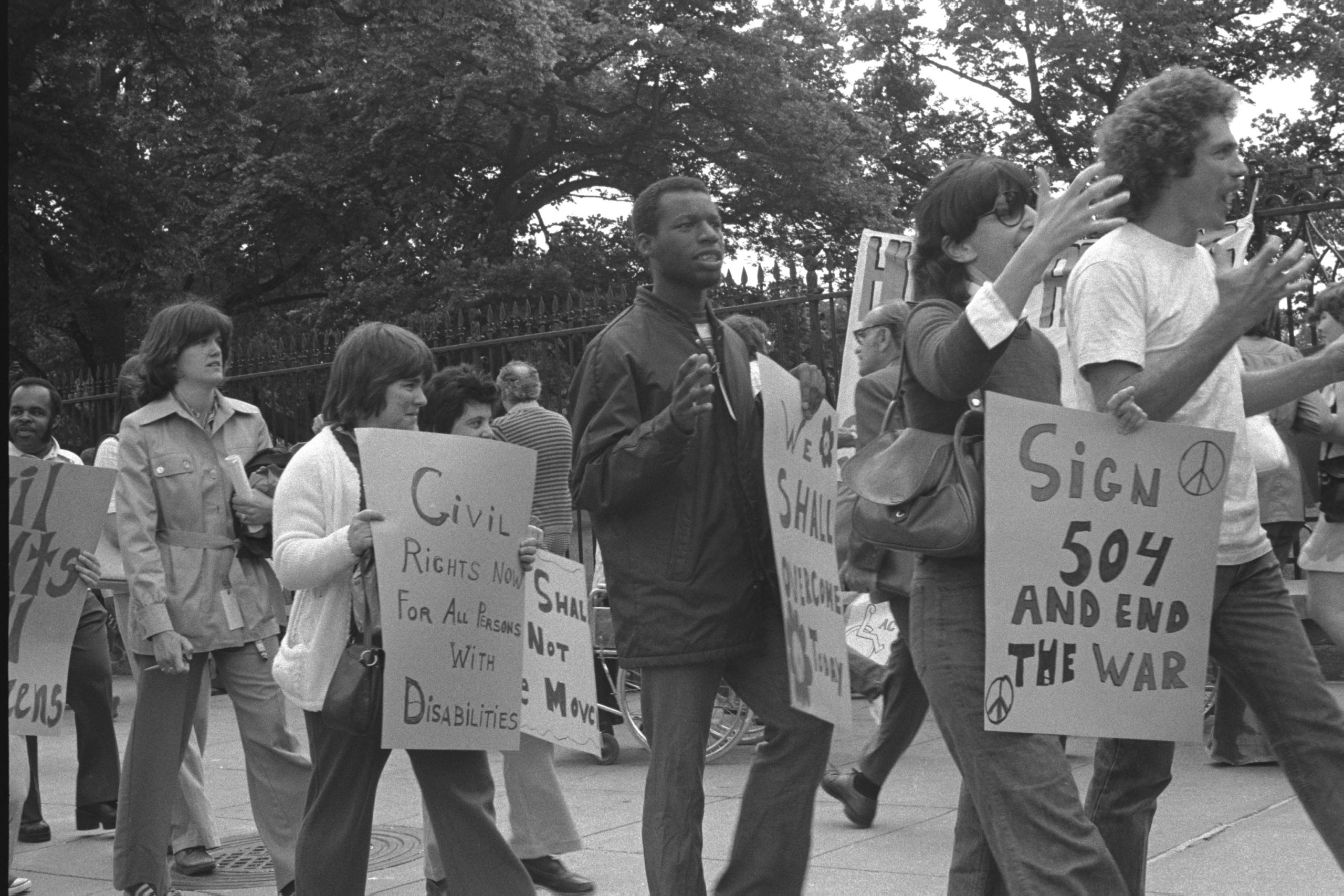 Personas manifestándose con carteles en lucha por los derechos de las personas con discapacidad para la toma de favor de la sección 504 en Estados Unidos. Las personas están manifestandose pacificamente con carteles que dicen: Derechos civiles ahora para todas las personas con discacapcidad, sección 504 y fin de la guerra...