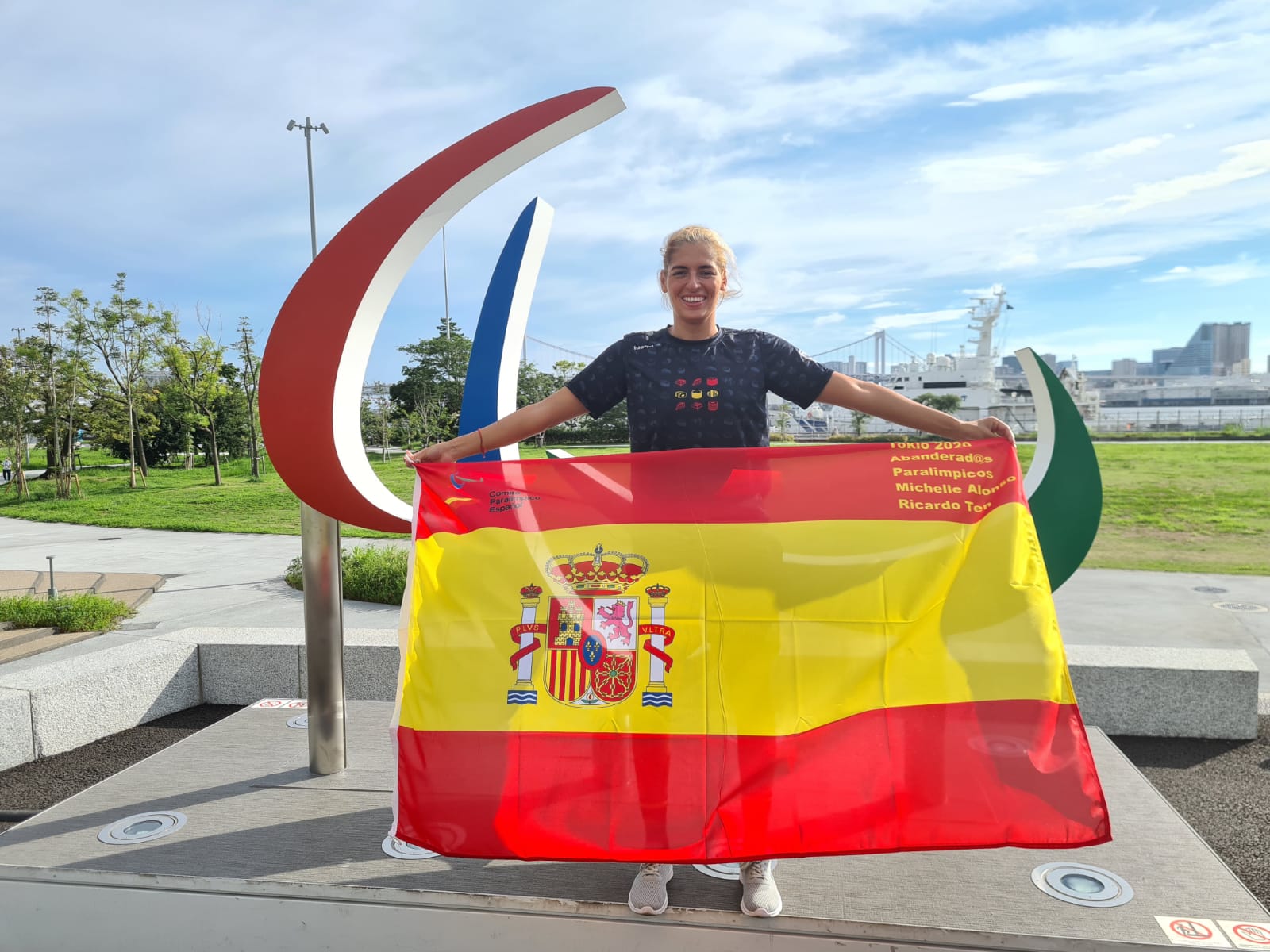 Michelle Alonso en Tokio posa frente al símbolo paralímpico mientras sonríe mostrando la bandera de España.