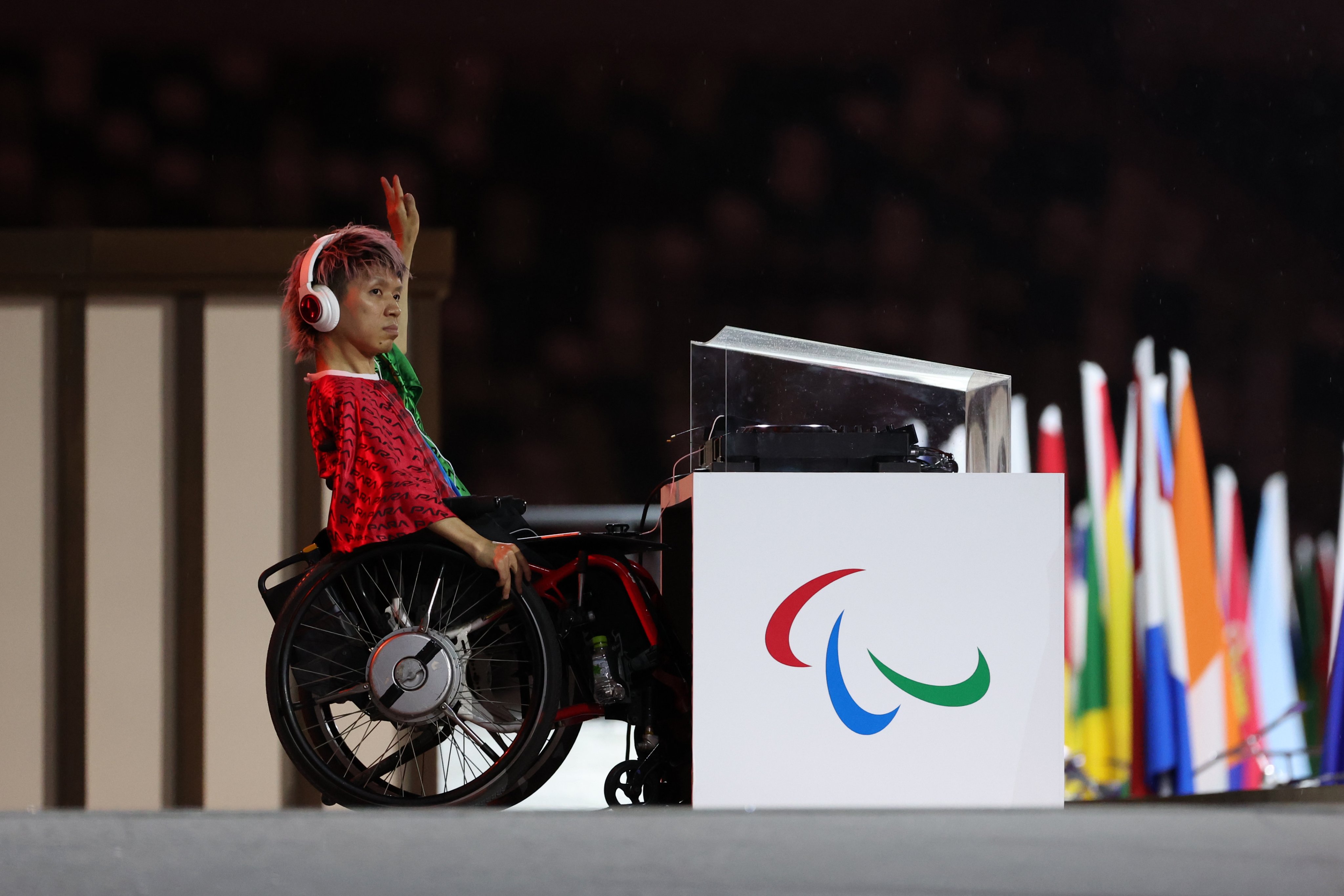 El dj Keita Tokunaga en la ceremonia inaugural. Aparece en su silla de ruedas y tiene a su lado su equipo de dj. Está levantando la mano para saludar.