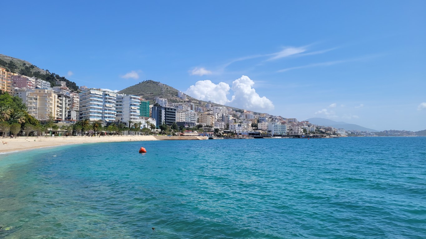 a view of the coastline in Saranda, Albania with blue water, yellow sand, and buildings lining the hills