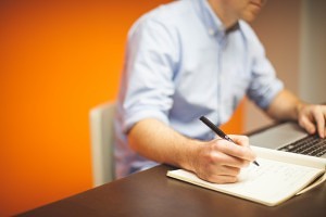a man writing on his book while looking at his laptop