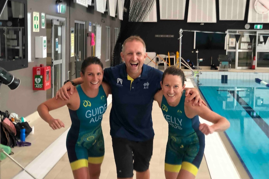 A para-triathlete smiles at the camera, standing next to her coach and her sighted guide.