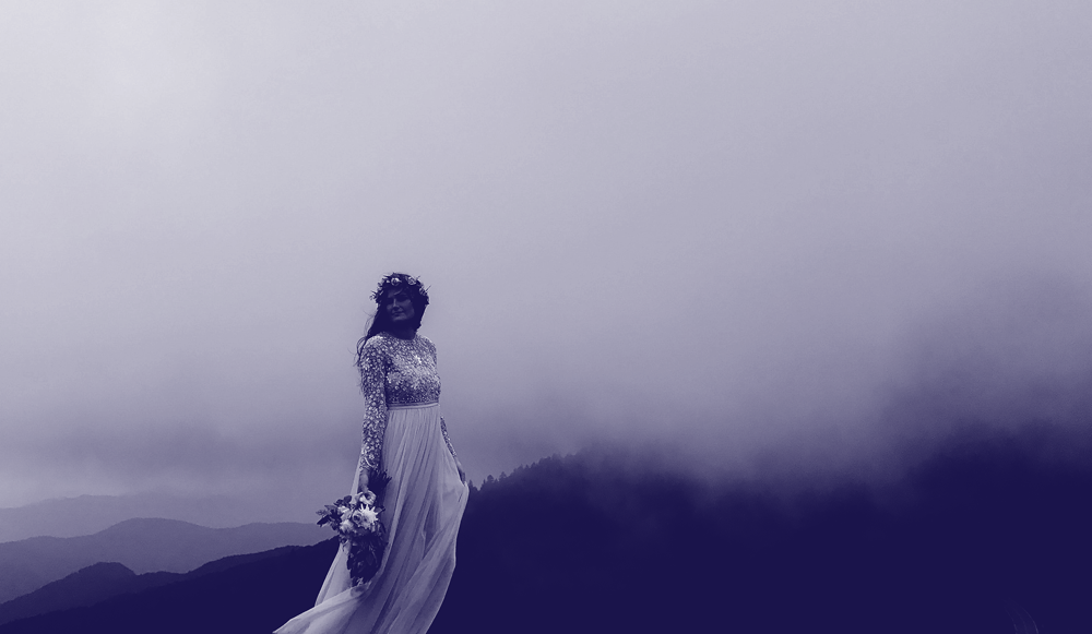 Image of a fair skinned dark haired woman in a white wedding gown standing against a vista of fog capped mountain ridges. The image has a dark purple overlay.