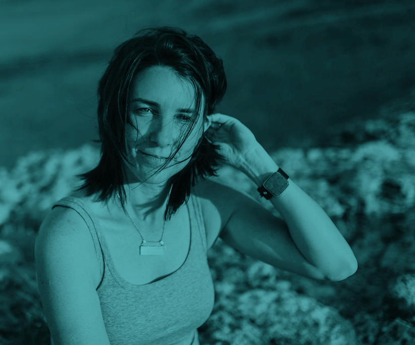 Pictured, Jenine Beecher - a fair skinned woman with shoulder length brown hair, she’s pushing some strands behind her left ear. She's wearing a neutral colored tank top and is pictured from the waist up. The image has a greenish tint.