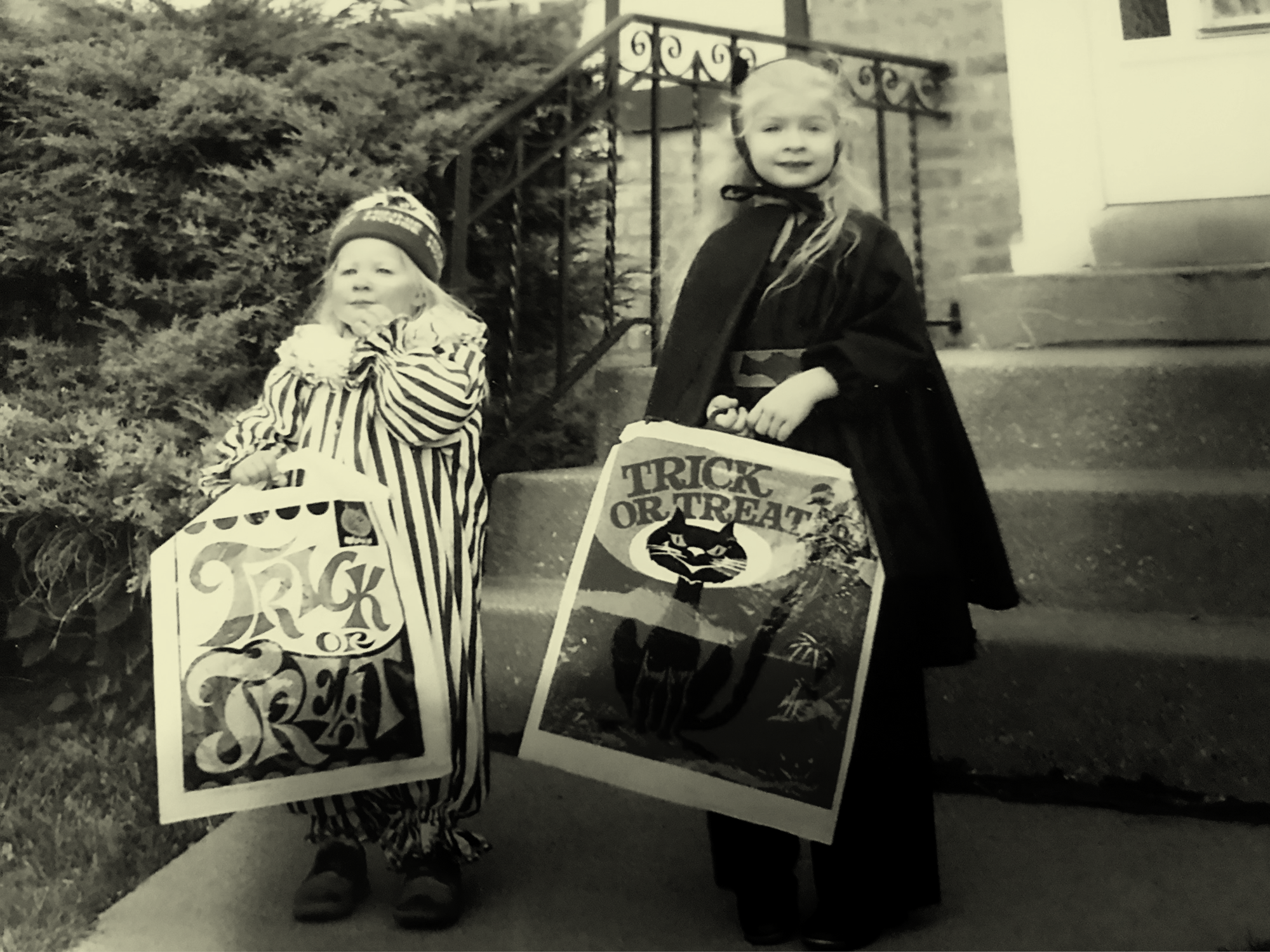 Two blonde and fair skinned children stand outside at the foot of a small cement staircase in homemade halloween costumes (a clown on the left, batgirl on the right), holding square plastic bags printed with halloween/trick-or-treat designs. The taller blonde child on the right (bat girl) is the author. The image has a yellow tint.