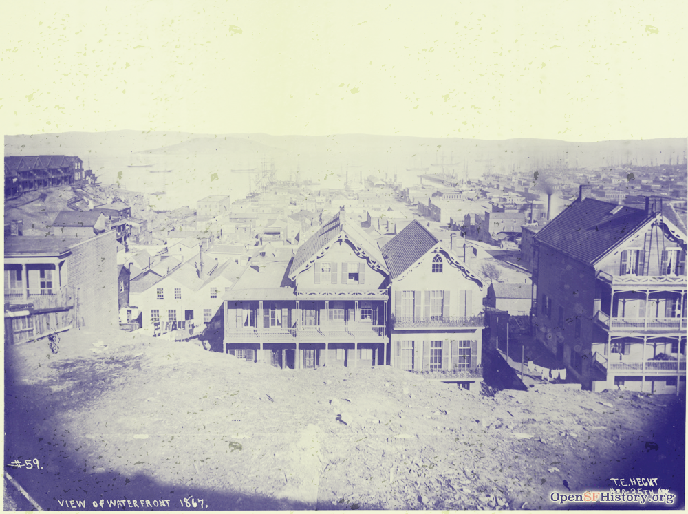 Photo of San Francisco in 1867 - the foreground is a barren dirt hill top looking out and down on wooden houses receding into the distance. In the far background you can see small ships floating on San Francisco Bay.s