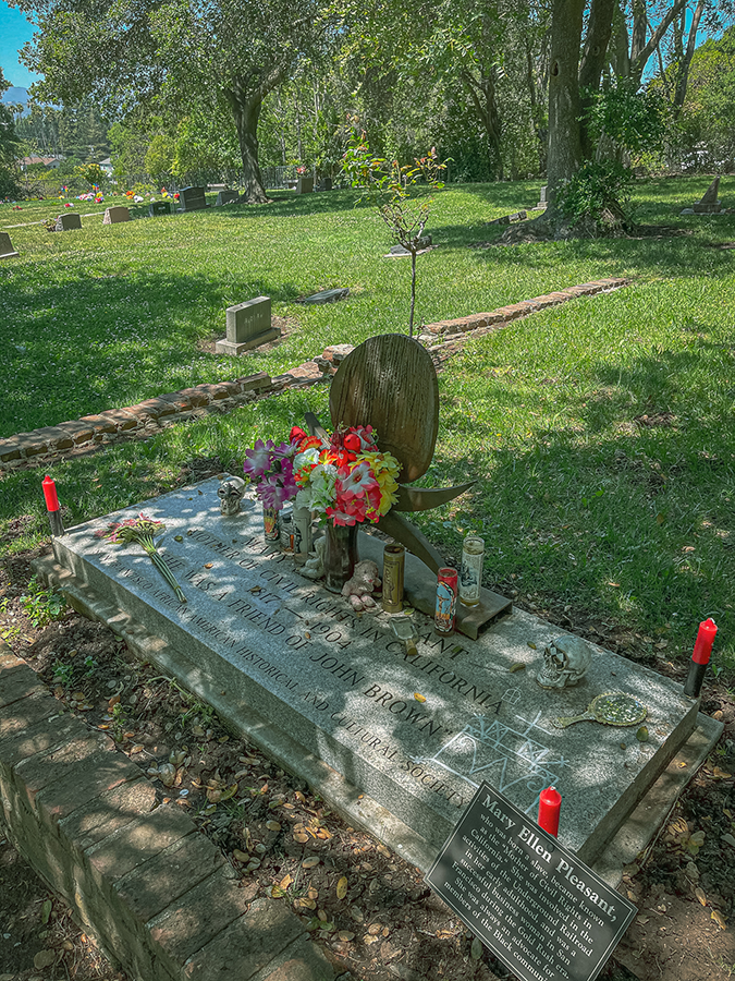 Photo of a grey stone grave marker with candles and flowers on top. The four corners of the marker have red candles. There are white markings drawn on the grave marker in white chalk or paint that look like a veve for Baron Samedi. The grave marker reads: “Mary Ellen Pleasant, Mother of Civil Rights in California, 1817-1904, She was a friend of John Brown, San Francisco African American Historical and Cultural Society.” There is a small sign on fixed to the grave corner that says: “Mary Ellen Pleasant, who was born a slave, blame known as the ‘Mother of Civil Rights in California.’ She was involved in the activities of the Underground Railroad in her early adulthood, and was a successful business woman in San Francisco during the Gold Rush era. She was always an advocate for members of the Black community.”