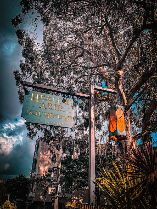 Photo of a street corner - there is a hanging sign that states "Healing Arts, 1801 Bush St." There's a hanging lantern oposite the sign. In the background there are mature eucalyptus trees. The photo has a very moody filter.