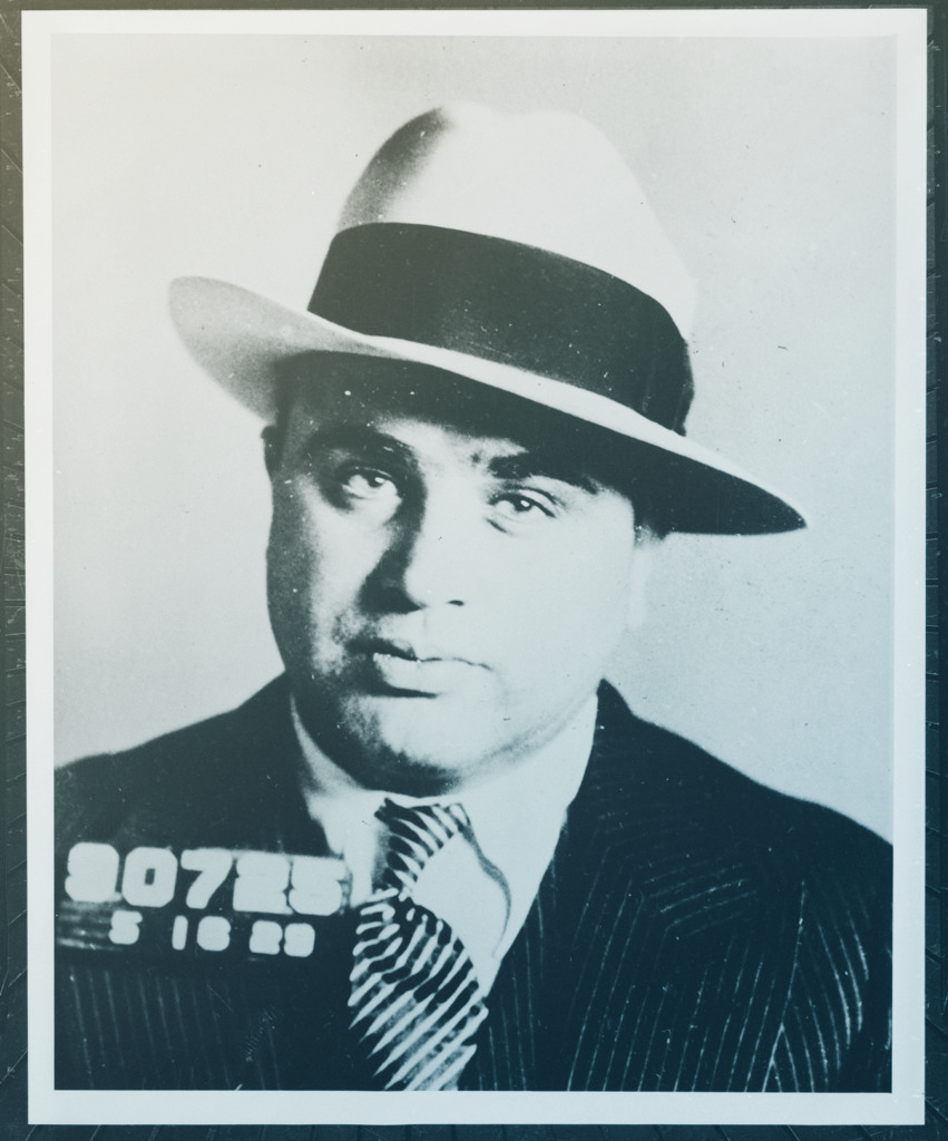 A mugshot of Al Capone. He's wearing a light colored fedora and a stripey suit and tie. He's wearing an expression that suggests he's not sorry, and this is all a bit of a hassle.