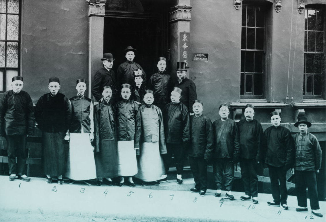A photo of an assembled group of Chinese employees and benefactors of the Tung Wah Dispensary arranged on the front steps of the building, circa 1900. There is a small plaque to the right of the doorway labeled Oriental Dispensary