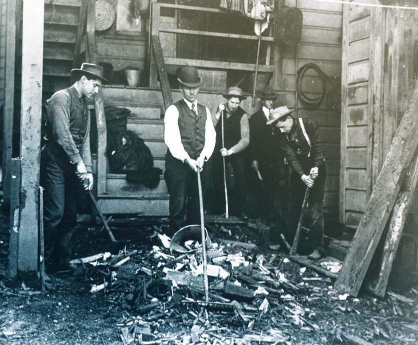 5 men in suite vests and hats all work with rakes in a semi-circle around a pile of burnt wood and paper debris