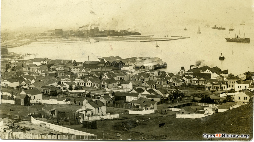 An aerial photo of clustered wooden buildings at the edge of the San Francisco bay. The buildings are a little haphazard, and the streets are bare dirt
