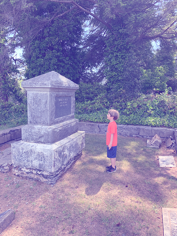 A young boy stands on front of a large stone grave monument – the monument is twice the height of the small boy