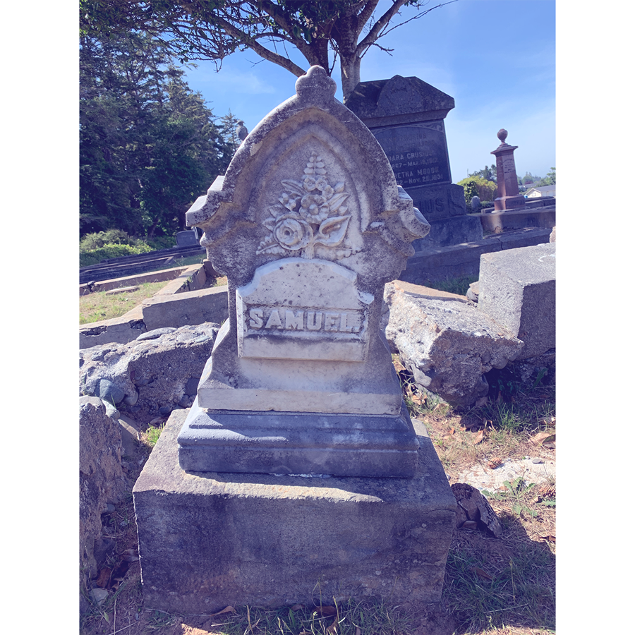 A tilting grave monument showing a pointed top, an etching of a floral bouquet, above large letters spelling out "Samuel." Any other marker information is lost or was never included.