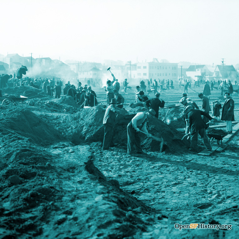 Many men working with shovels and hammers in a dirt field amidst great upturned piles of earth and filled wheelbarrows
