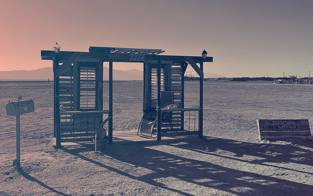 An art installation in the sand around Salton Sea, CA - A porch made of scrap wood and metal, with an awning and wooden shutters. There's a camp chair on the porch, and a sign above reads "777." In front of the scene is a mailbox standing upright in the sand. To the right of the scene is a low sign that reads, "The Porch View." In the distance behind you can see the edge of the water.