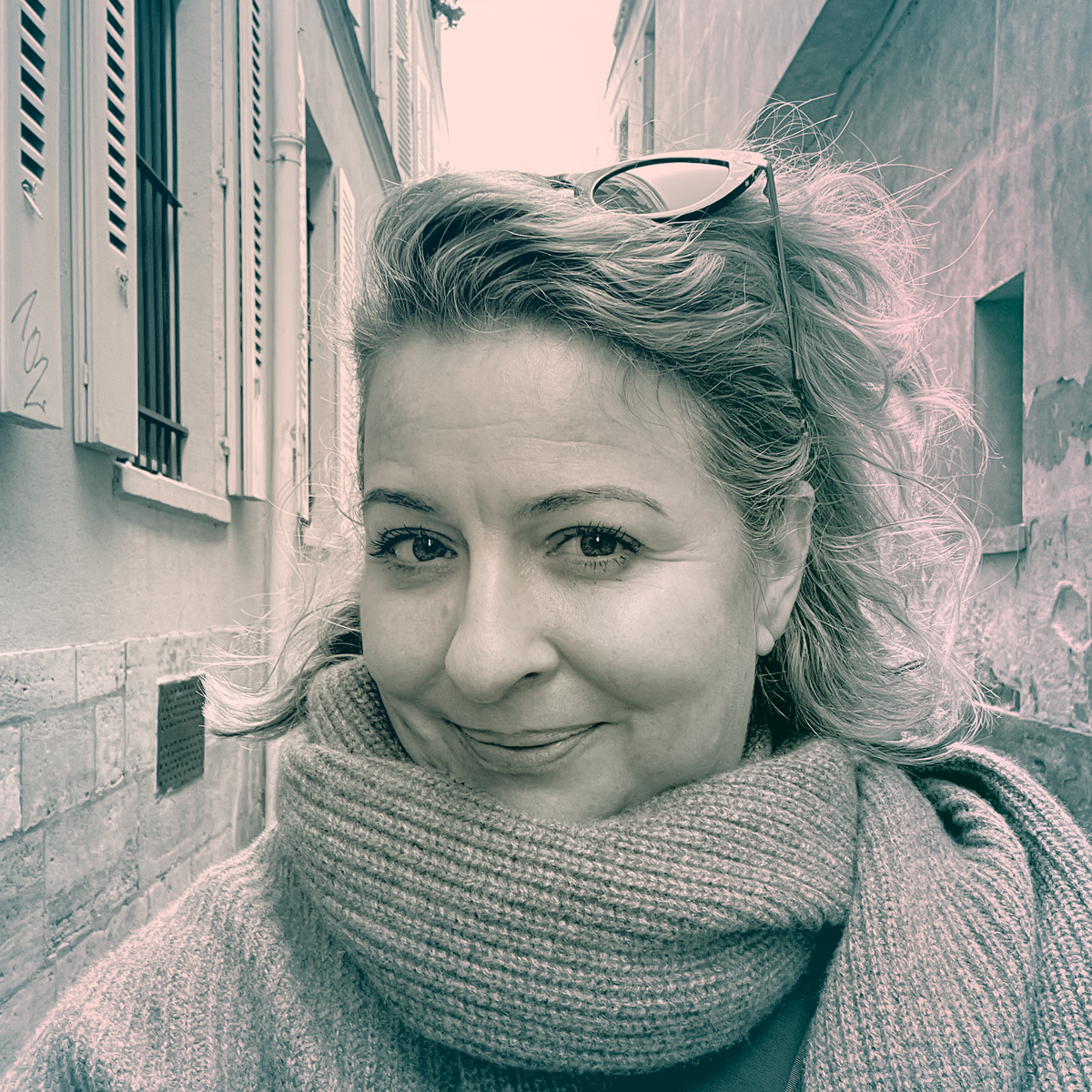 A headshot of Courtney , hair pushed back and held with a pair of sunglasses on top of her head, a thick scarf around her neck. In the background the tall stone walls of the Rue des Chantres.