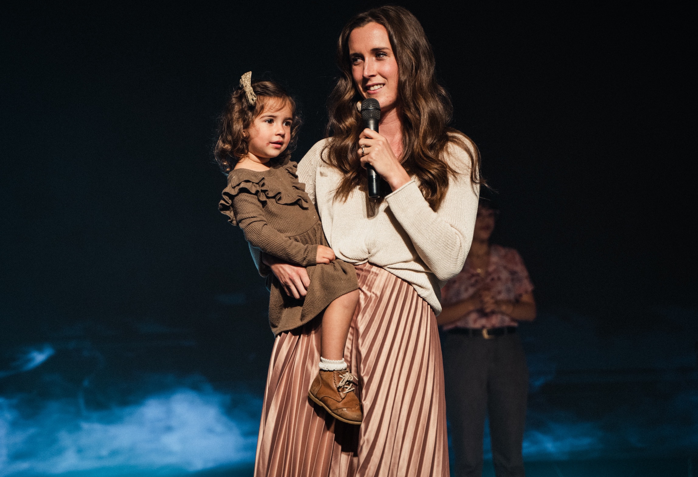 Woman with girl toddler on her hip talking into a microphone