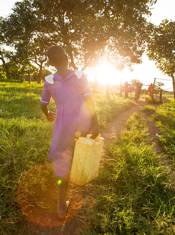 Girl with back towards us, walking towards sunset while carrying water container. 