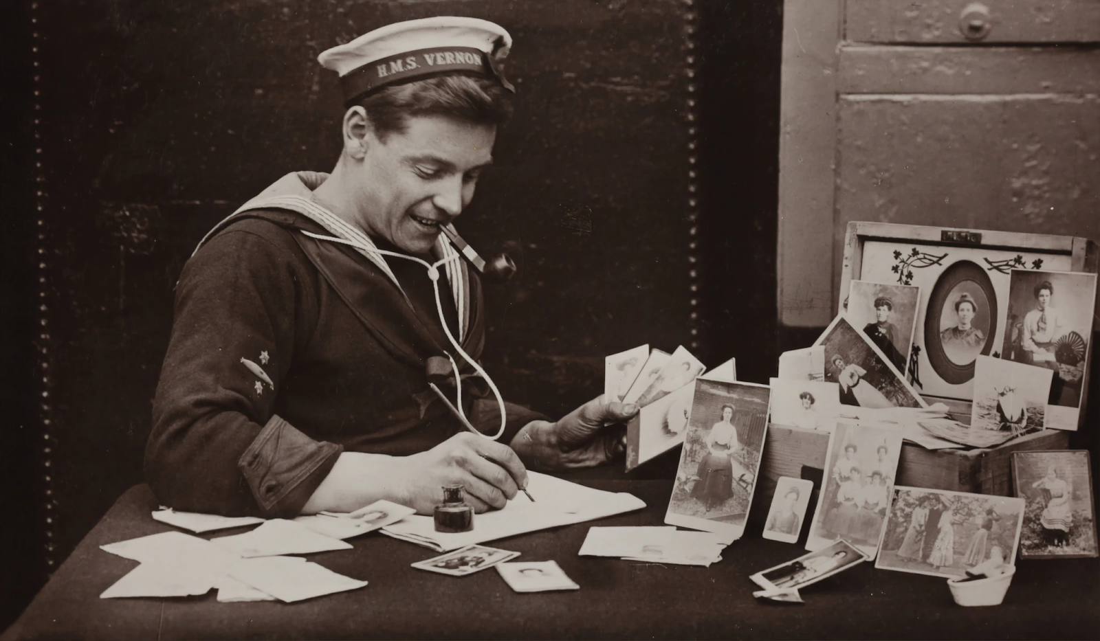 A black and white image of a old-fashioned marine writing a letter. He is surrounded by pictures of a beautiful woman.
