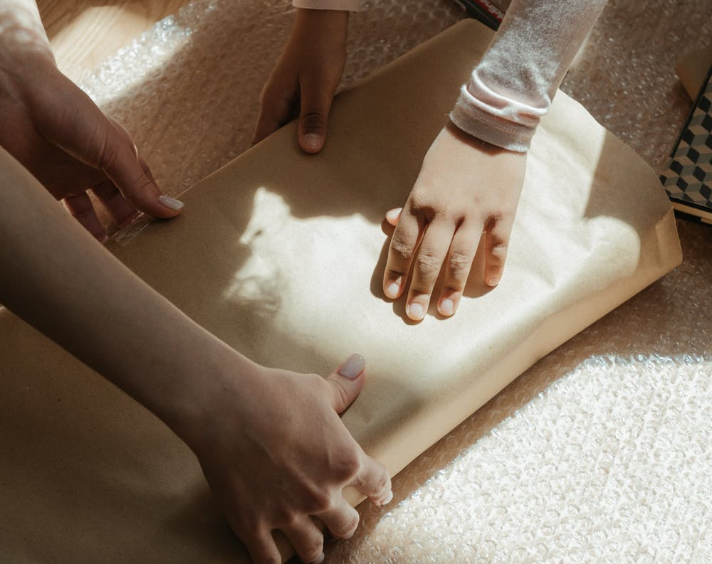 An image of the hands of both a woman and child wrapping a box in paper and bubble wrap.