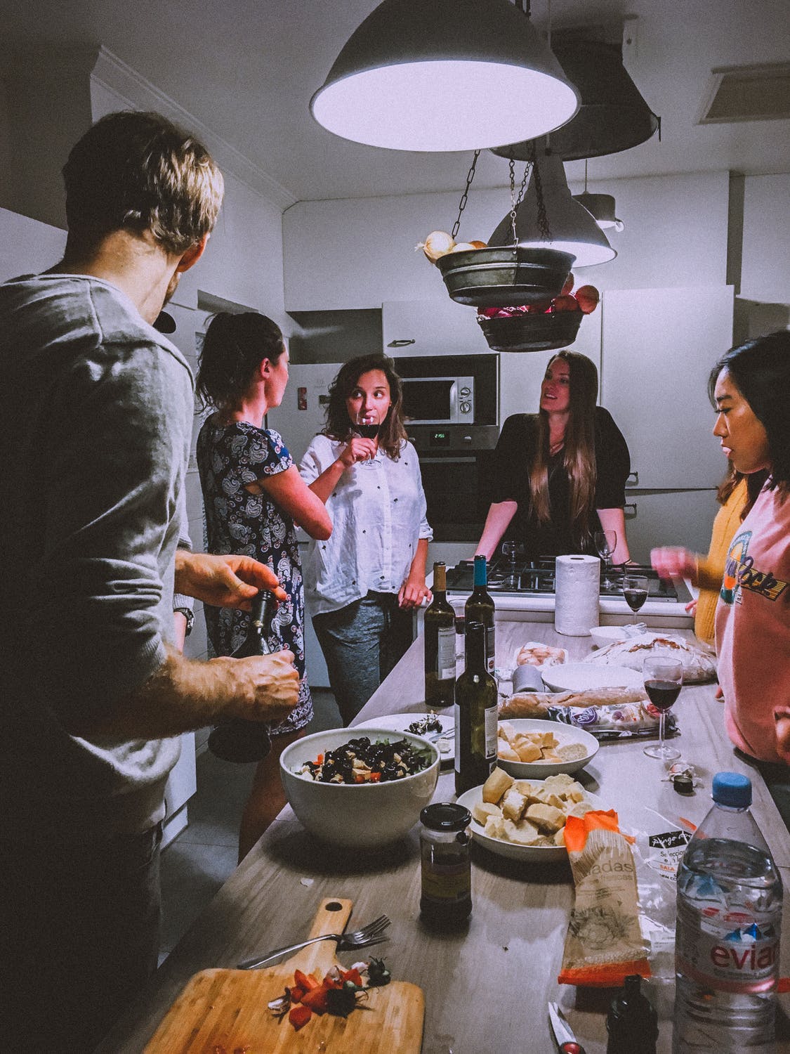 Group of friends stand around a kitchen island drinking wine and eating snacks