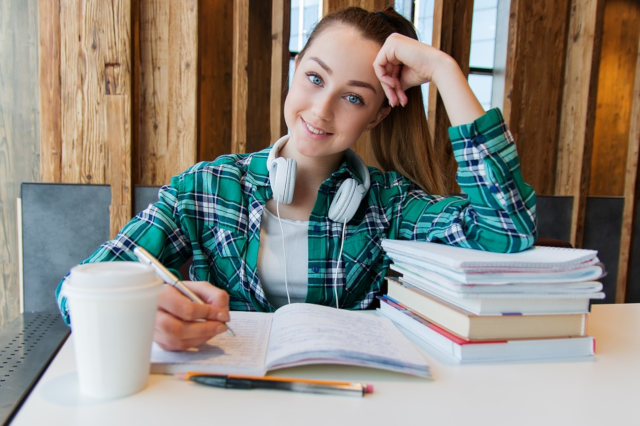 college student studying, girl with pony tail writing in book at table with arm braced on pile of books, wearing denim jacket with headphones