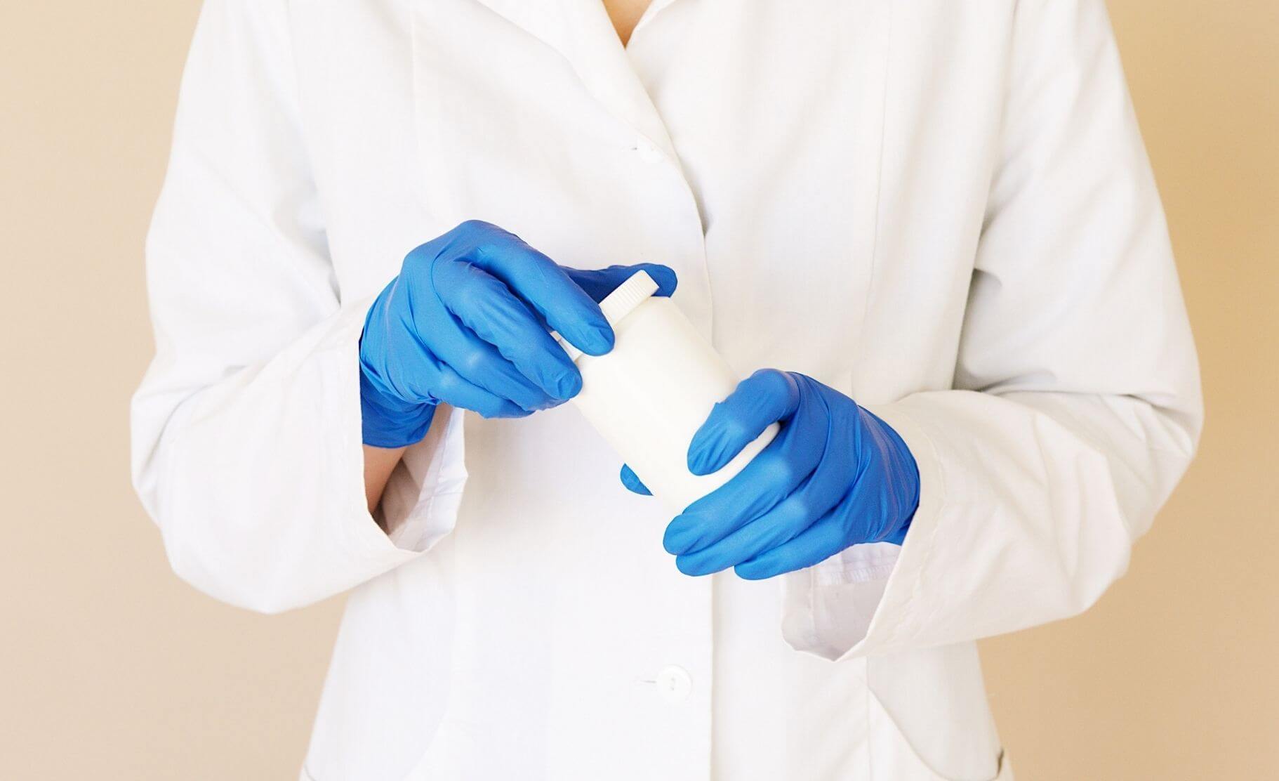 A doctor in a white lab coat holds a large, white pill bottle with hands covered in disposable sanitary gloves