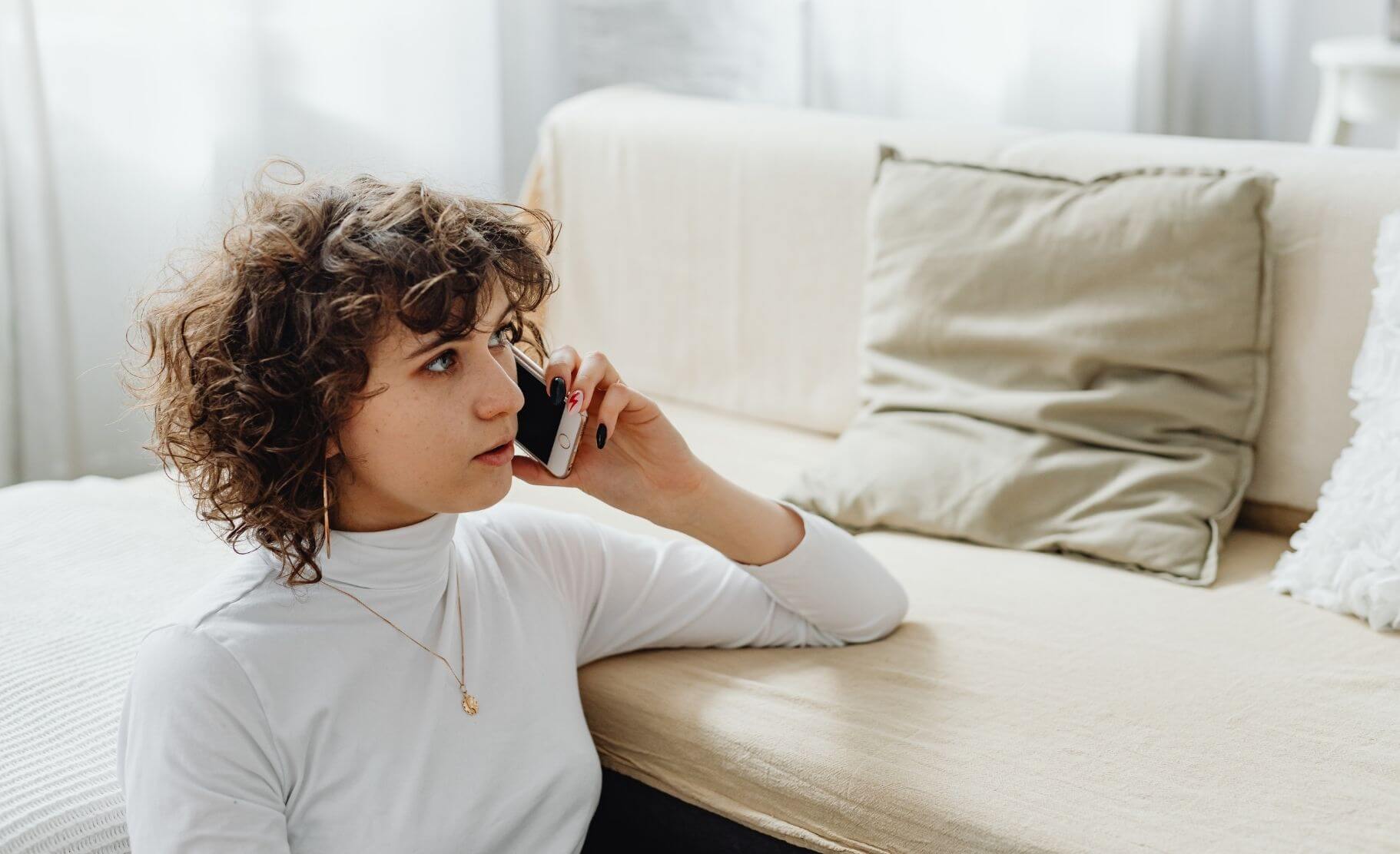 A woman siting on the floor next to a couch in a bright room speaks on the phone