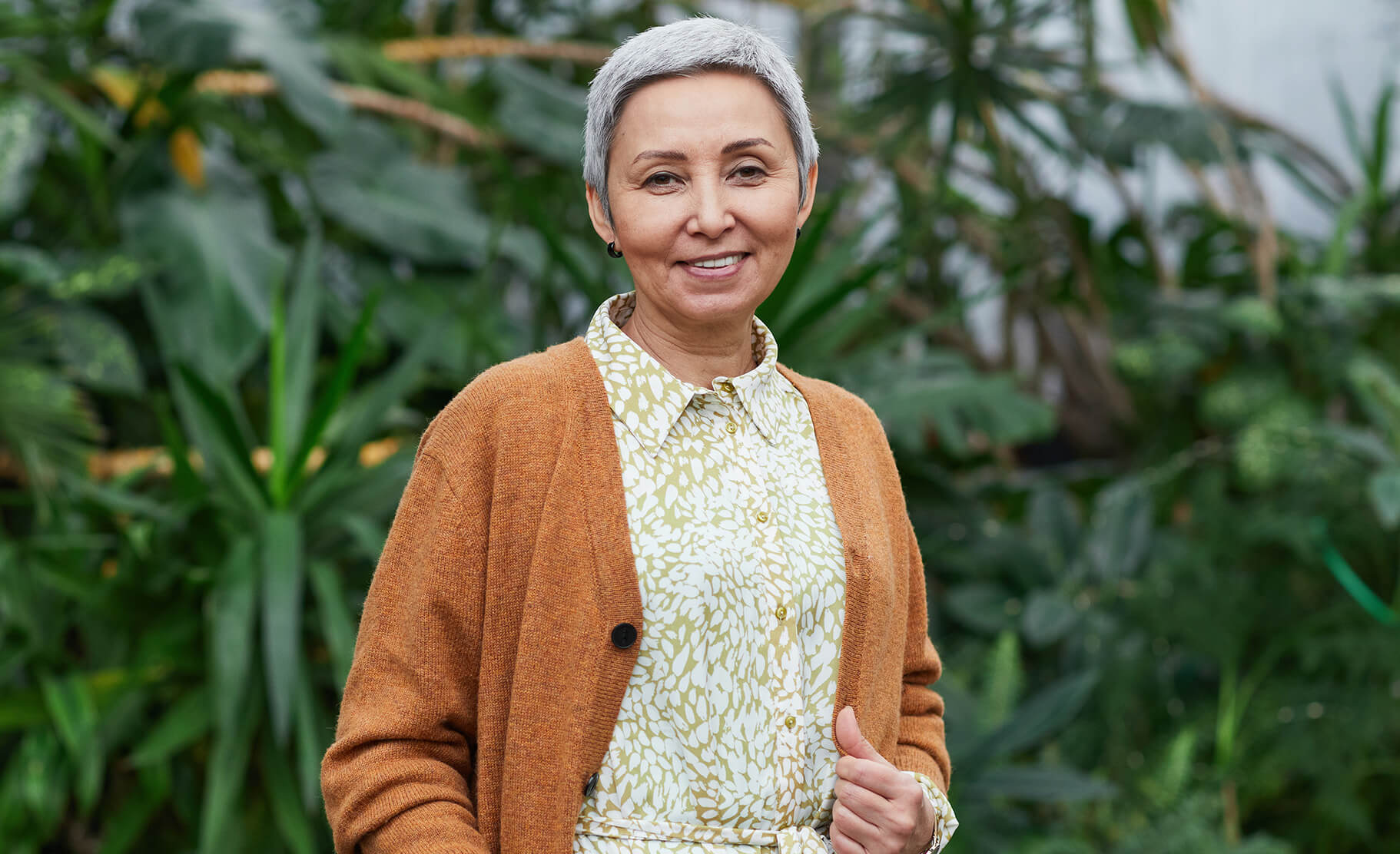 A grey-haired woman in an orange cardigan poses for a portrait in front of green plants