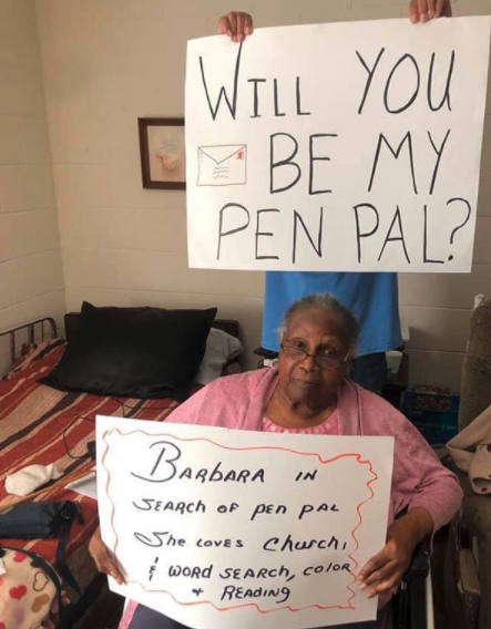 a nursing home resident holds a sign saying "will you be my pen pal?"