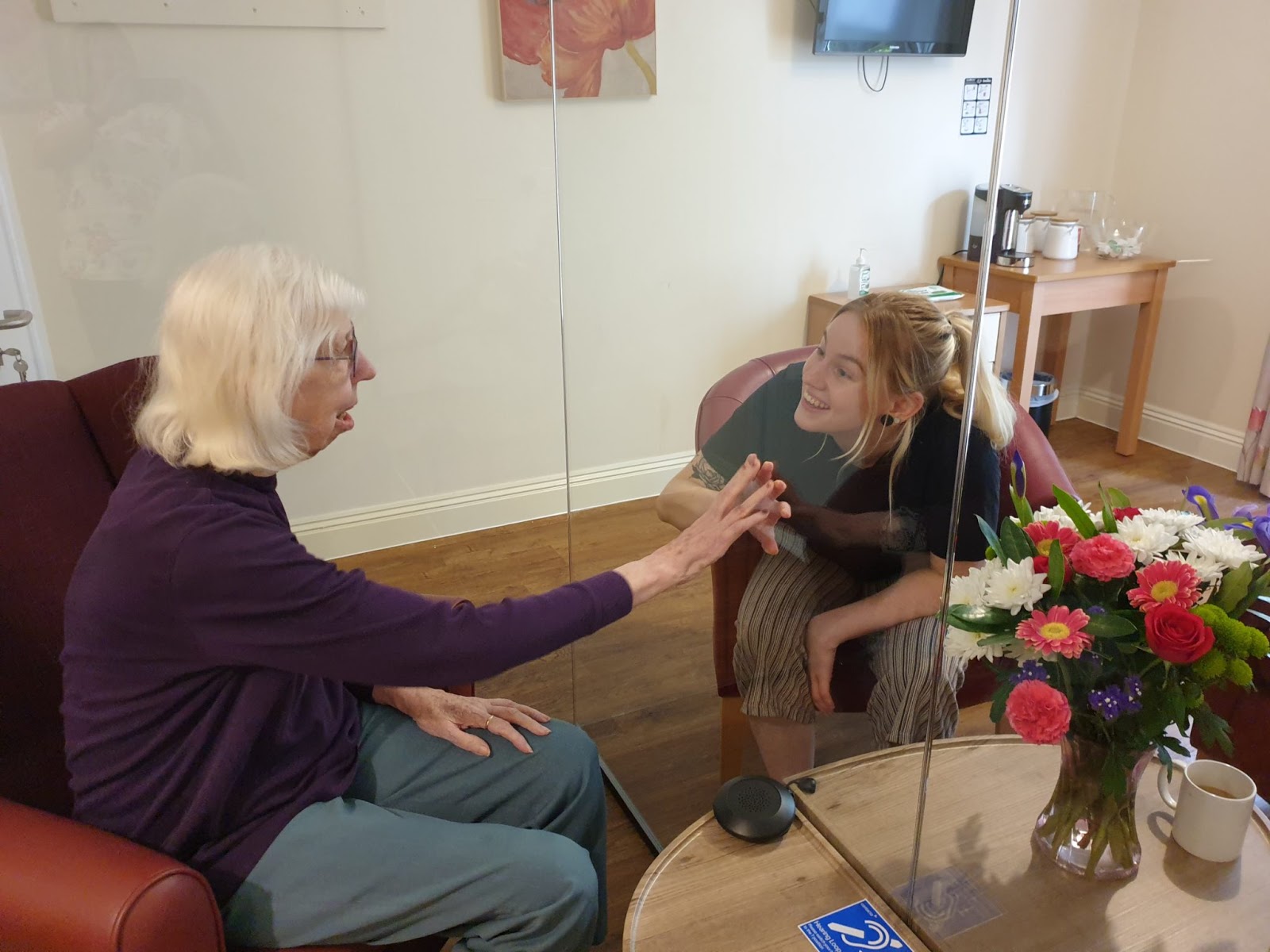 a care home resident greets her visitor through a glass partition