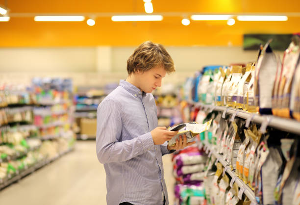 Man wearing blue collared shirt reading product information and also using smartphone in pet food aisle at a pet sotre