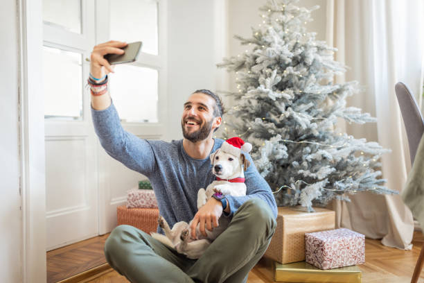 Our favorite time of the year Young man playing on floor with his dog. Christmas three in background. Dog is wearing Santa's hat decoration on head. dog christams camera stock pictures, royalty-free photos & images