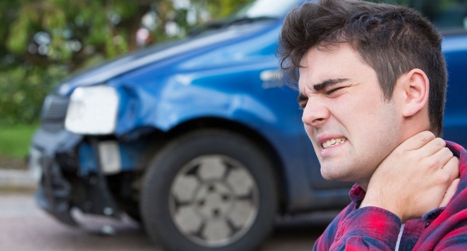 The driver of a vehicle has a pained face while standing near their smashed car and holding their neck.