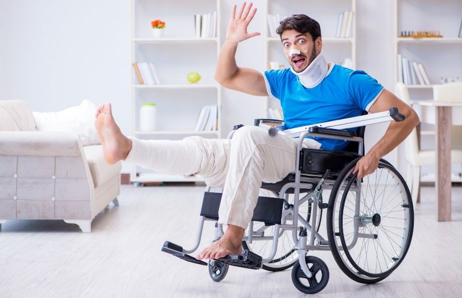 A person with a neck brace and cast leg in a cast sits in a wheelchair in his living room and waves to you.