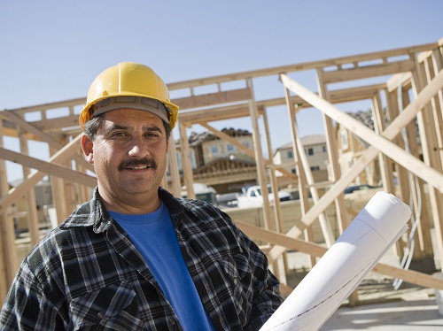 Construction worker at outdoor construction site holding contractor plans and wearing a yellow hardhat.