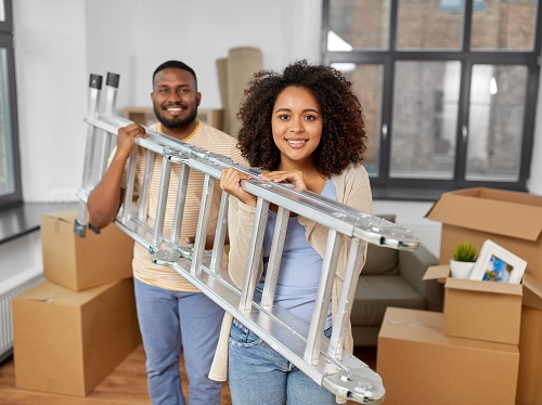 A man and a woman carrying a ladder together inside a home.