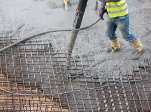 Worker wearing boots and gloves pouring concrete onto rebar formwork.