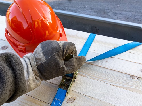 Worker wearing a glove is tightening a strap that is securing lumber on a trailer and a red hardhat is nearby.