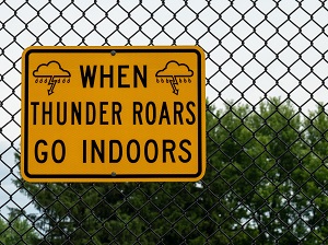 A chain link fence with a yellow and black sign hanging on it that says, "When Thunder Roars Go Indoors."